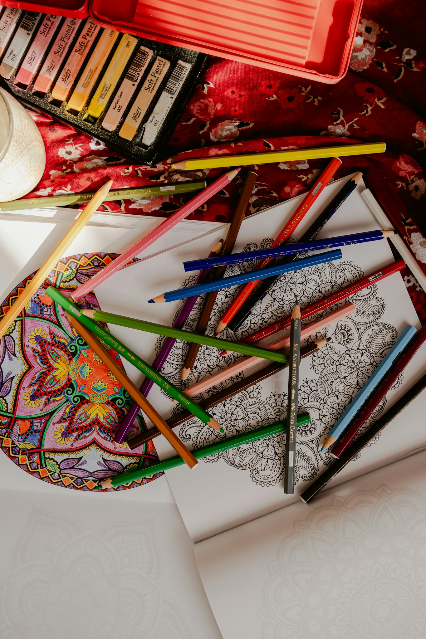 Vibrant mandala with colored pencils on a floral red fabric, creating a lively artistic setup.