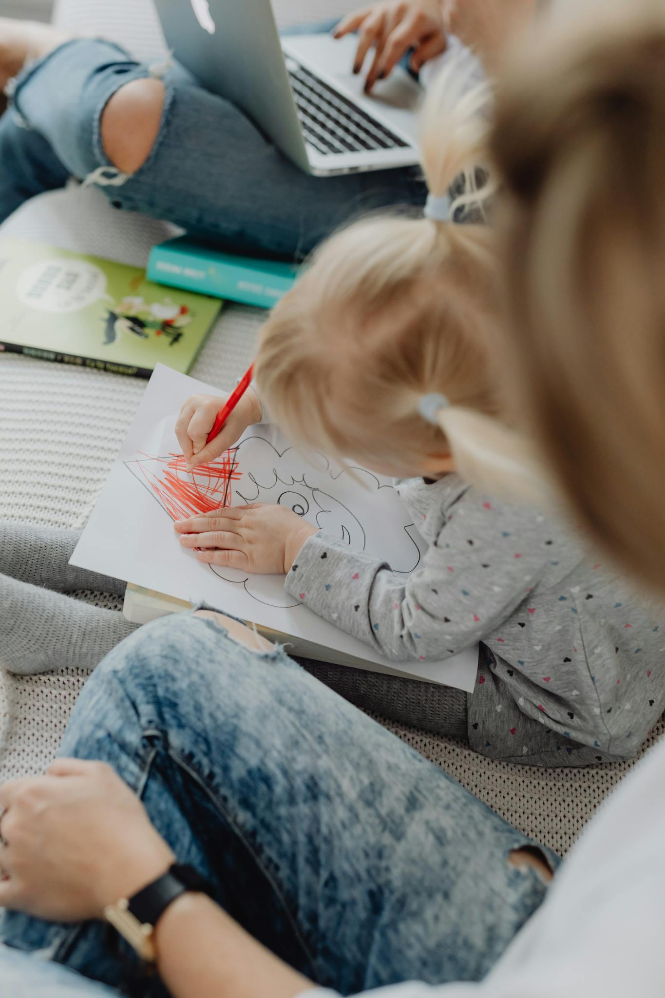 Young child coloring with a red crayon while under adult supervision in a cozy home setting.